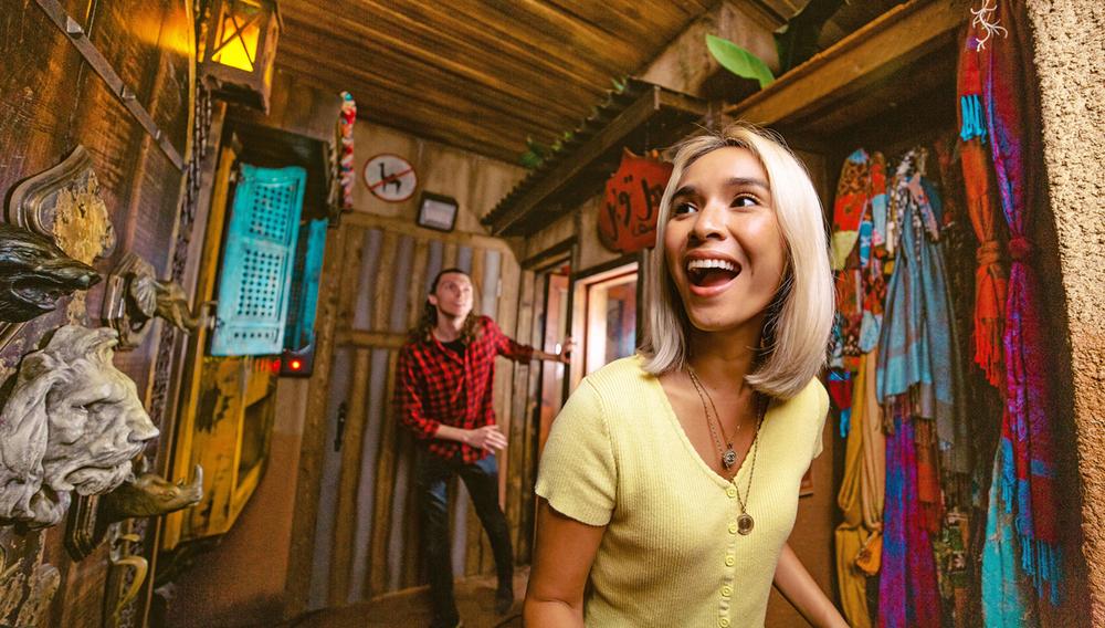Image of 2 escape rooms players exploring a room that is decorated with colorful fabrics hanging on one wall and a stone lion head on the left.